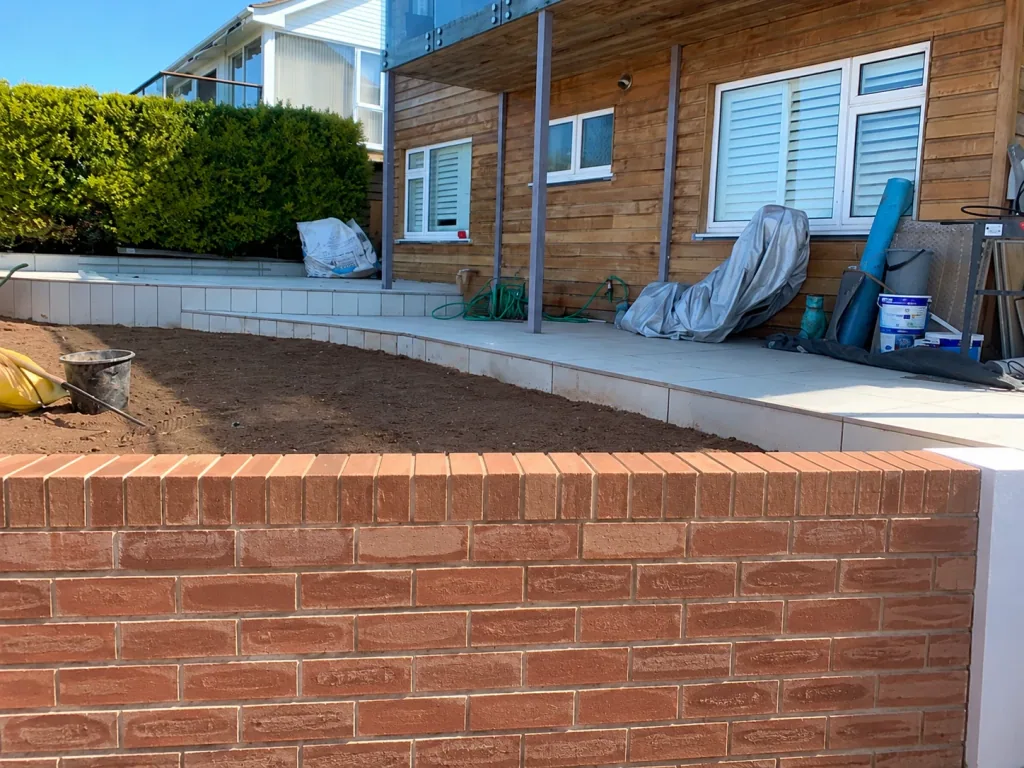 Work in progress at a timber-clad property: a new red brick retaining wall with a soldier course, prepared ground behind it, and pale paving already laid along the building.